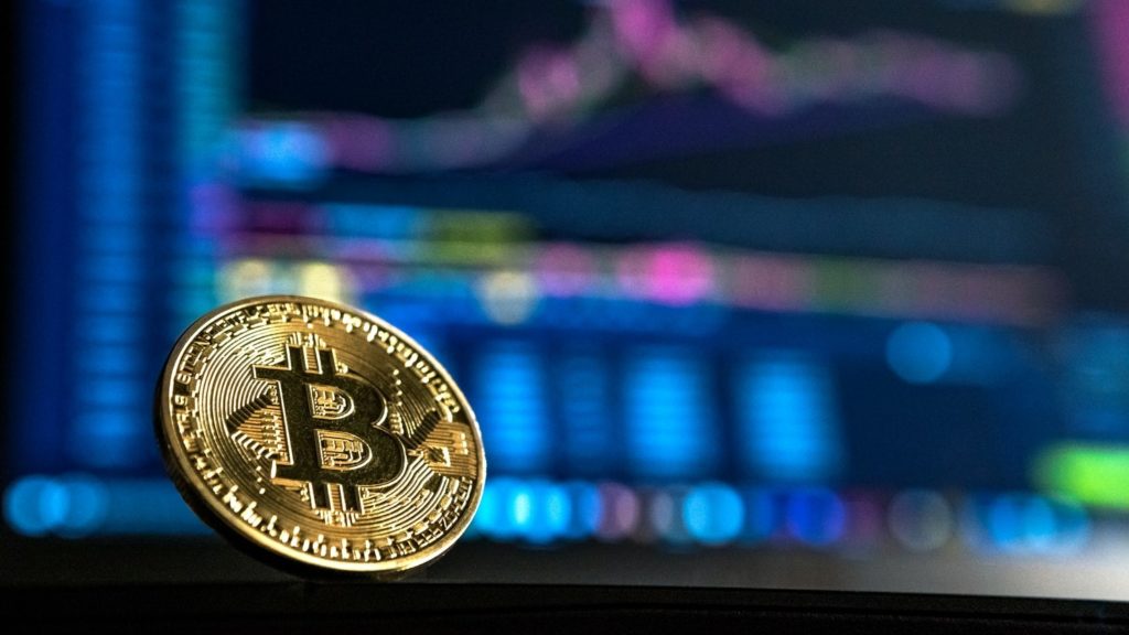 Imagen de una moneda de Bitcoin en posición vertical descansando sobre un fondo gráfico.