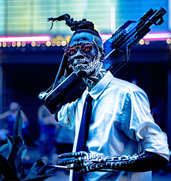 Una imagen de un cyberpunk caminando por una calle colorida, su cuerpo pintado para imitar una máquina, mientras porta un arma automática.