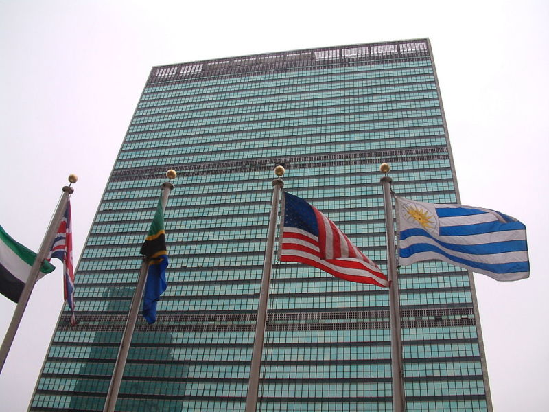 Rascacielos de vidrio y acero con banderas de varias naciones frente a él.