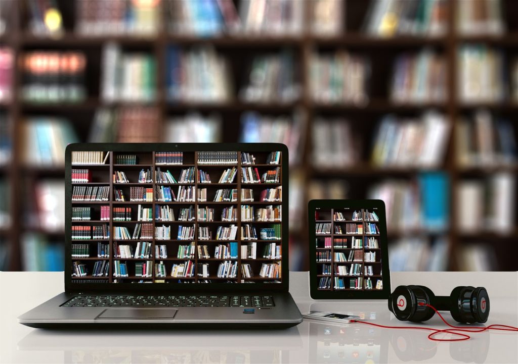 Imagen de una computadora portátil y una tableta en la mesa frente a una estantería llena de libros. Las pantallas de las computadoras portátiles y las tabletas muestran una imagen de una estantería llena de libros.