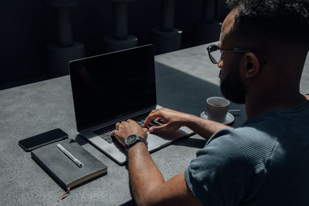 Imagen de un hombre escribiendo en una computadora portátil con la libreta a la izquierda y la taza a la derecha.